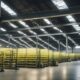 Harsh fluorescent light floods the cavernous warehouse, casting stark shadows on towering shelves and machinery